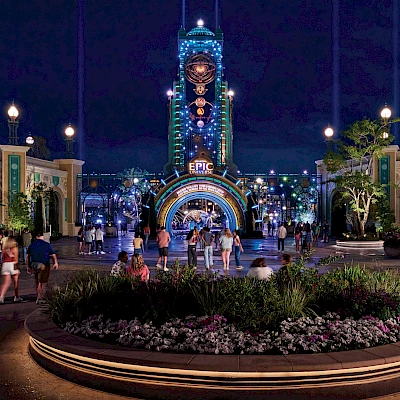 Nighttime scene of a theme park entrance, featuring colorful lights, a tall clock tower, and a crowd of people gathered around a garden.
