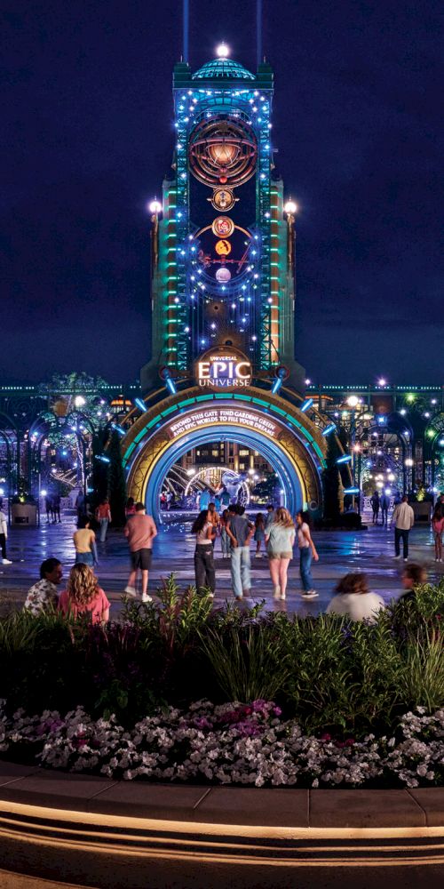 Nighttime scene of a theme park entrance, featuring colorful lights, a tall clock tower, and a crowd of people gathered around a garden.