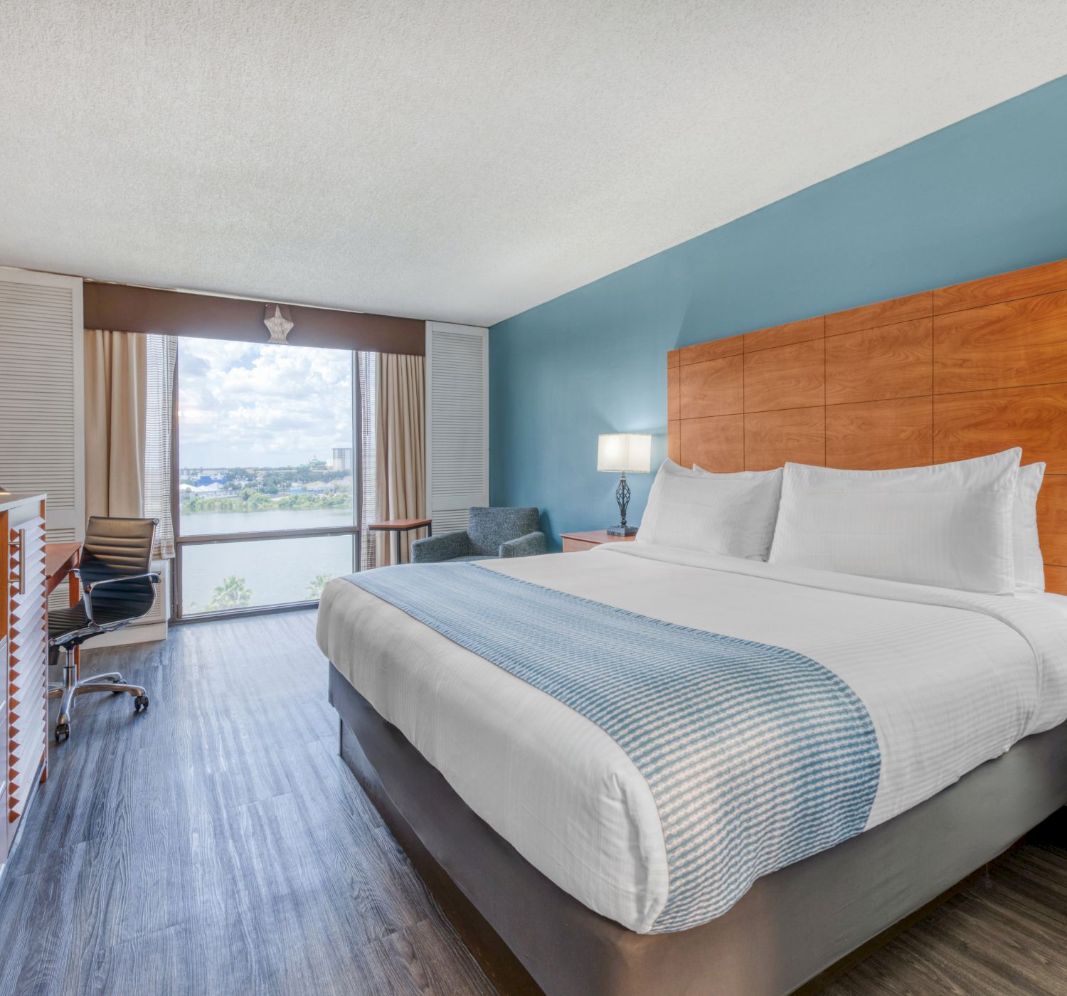The image shows a modern hotel room with a large bed, bedside lamps, a TV, desk, and a window with a view.