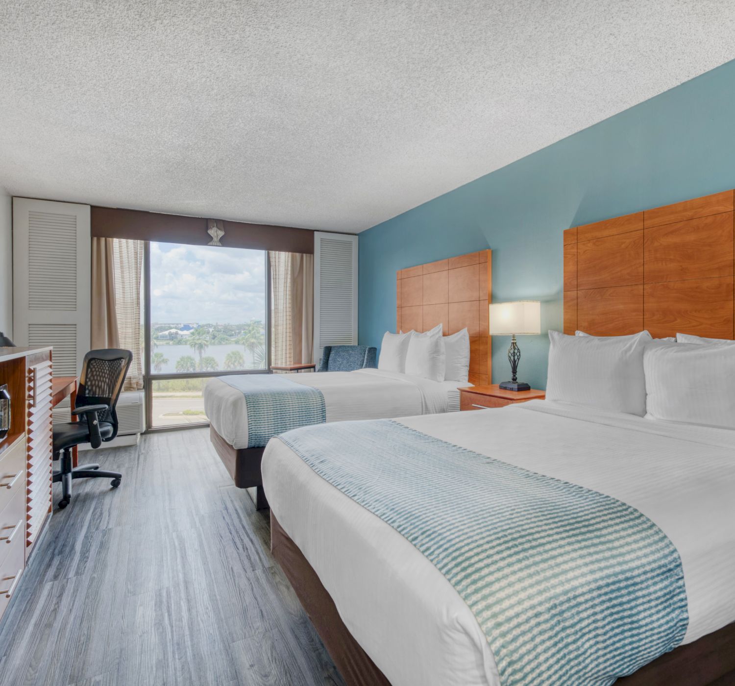 The image shows a hotel room with two beds, a TV, a desk and chair, and a window with a view. The decor is modern and minimalistic.