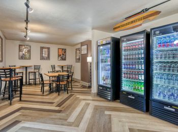 The image shows a room with tables, chairs, and beverage coolers filled with drinks. Posters decorate the walls for a cozy vibe.