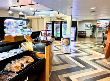 A store interior with wooden flooring, stocked shelves, a counter with pastries, and refrigerated beverage displays in the background.