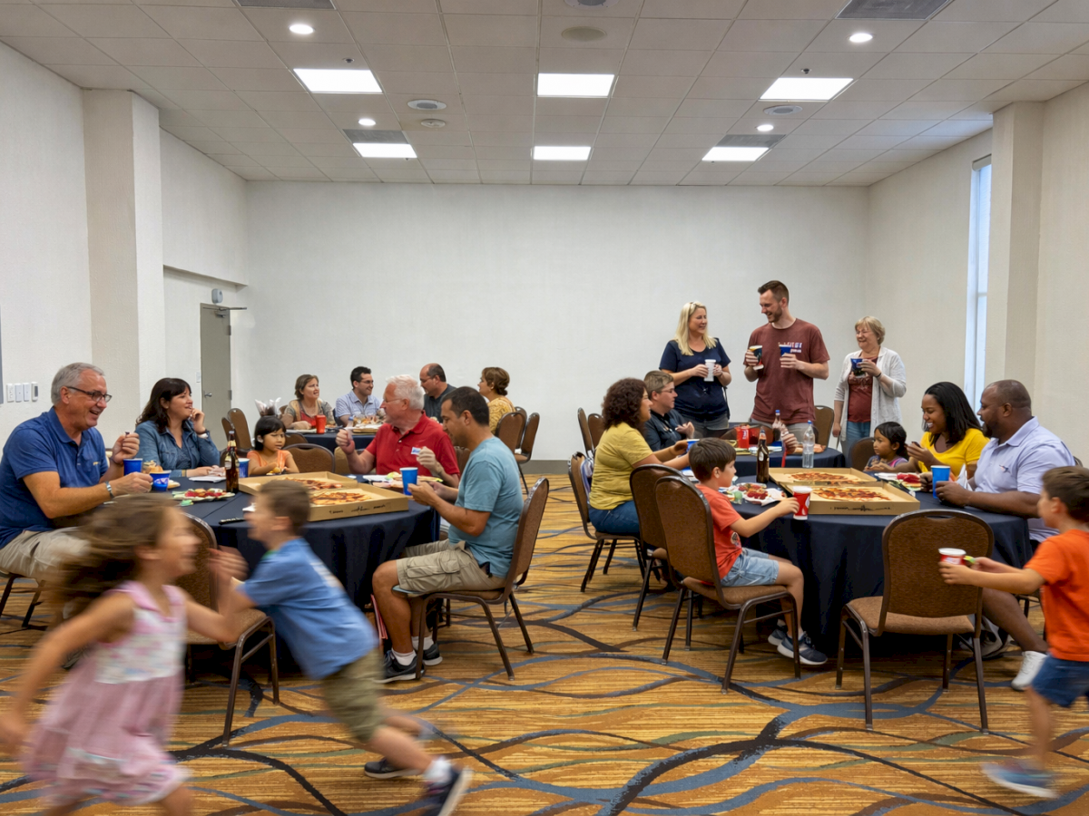 A group of people sits at tables in a room, dining and socializing, while children play around them in motion.
