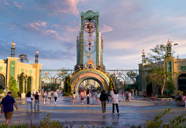 The image shows people walking through a vibrant, themed entrance with an arch and tower, under a colorful sky.