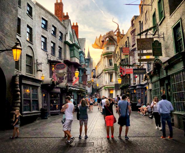 People walking through a themed street with a dragon sculpture above, among various shops and medieval-style buildings, under a partly cloudy sky.