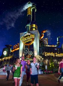 The image shows a whimsical, steampunk-style chocolate factory at night with people walking in the foreground.