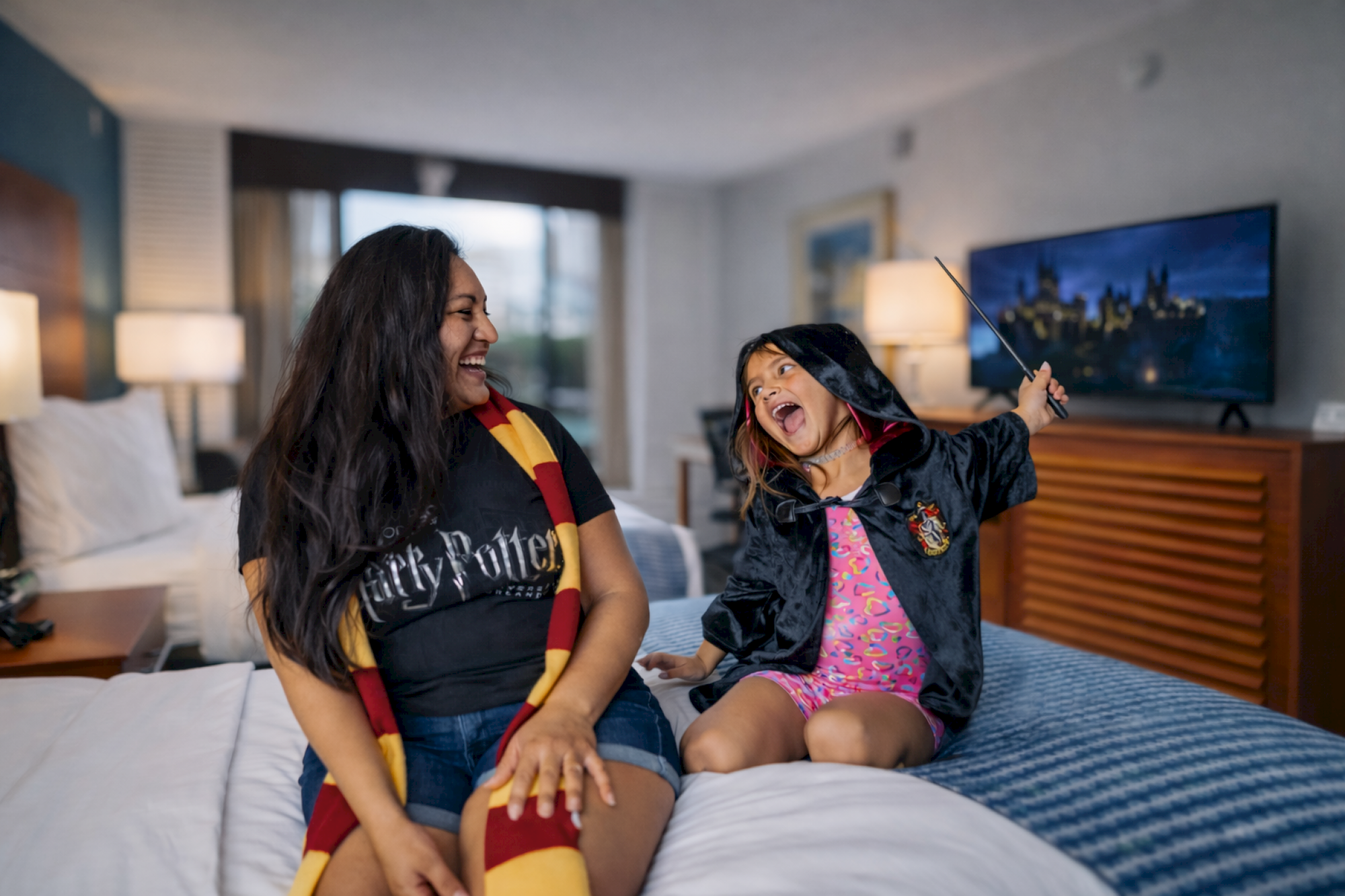 A person and a child dressed in Harry Potter attire are joyfully playing on a bed in a hotel room.