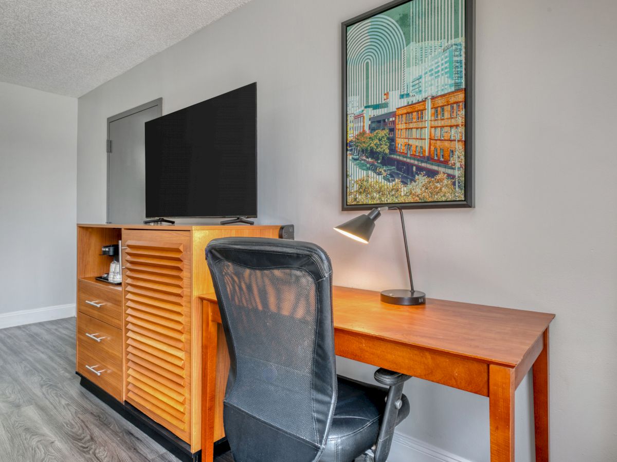 The image shows a workspace with a wooden desk, a rolling chair, a lamp, a TV on a cabinet, and framed wall art.