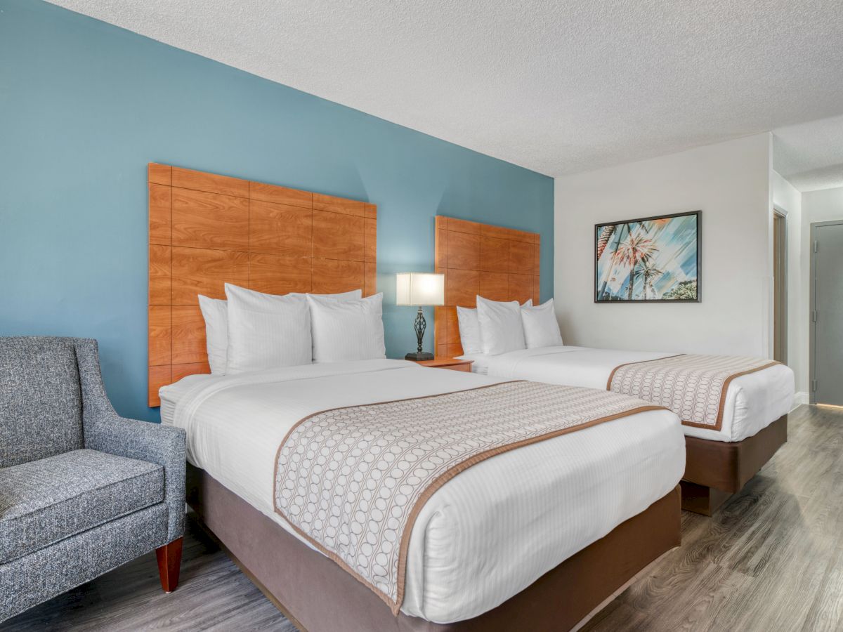 The image shows a hotel room with two double beds, a gray armchair, a nightstand with a lamp, and a wall painting, ending the sentence.