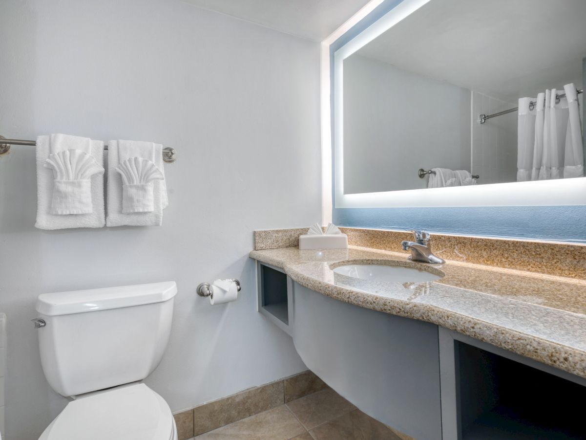A clean bathroom with a granite countertop, illuminated mirror, sink, toilet, and folded towels on a rack.