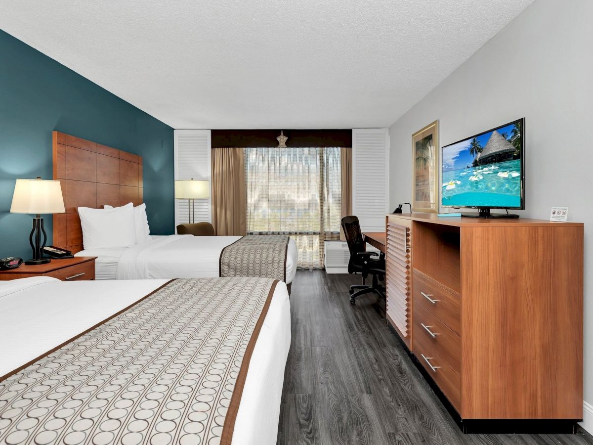 The image shows a hotel room with two beds, a TV on a wooden dresser, a desk with a chair, and a window with a curtain.