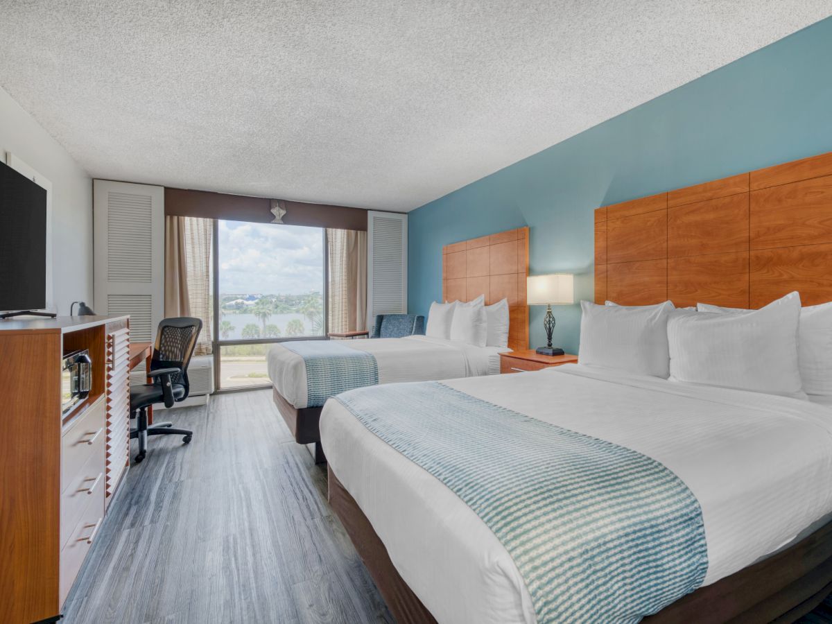 The image shows a hotel room with two beds, a TV, a desk chair, and a window with a view, featuring a light blue and wood theme.