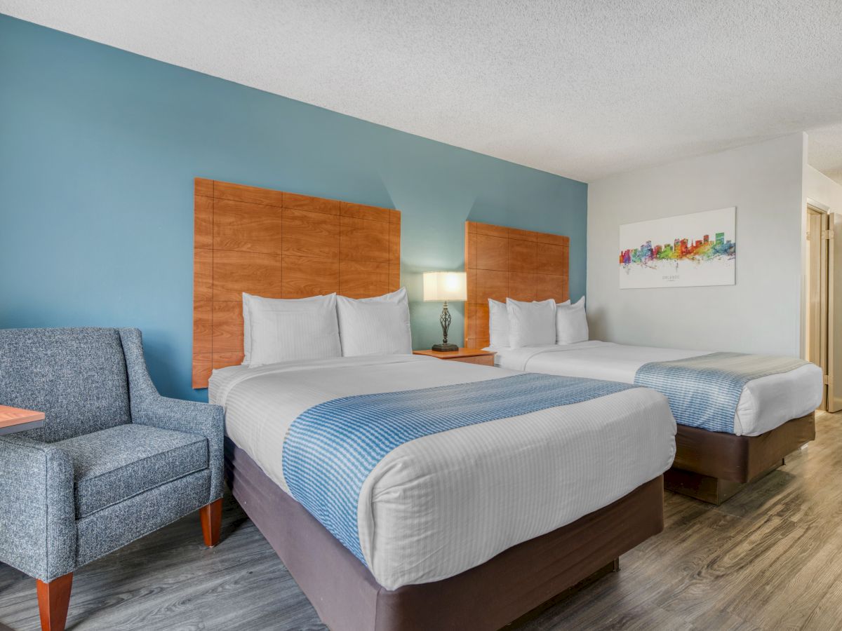 The image shows a hotel room with two beds, a blue accent wall, a gray armchair, and a colorful painting on the wall.