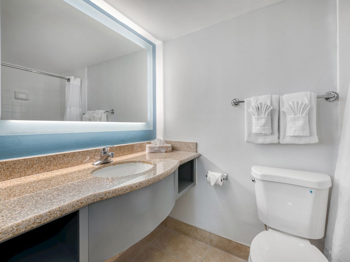 The image shows a modern bathroom with a sink, large mirror, toilet, and neatly folded towels on the wall rack.