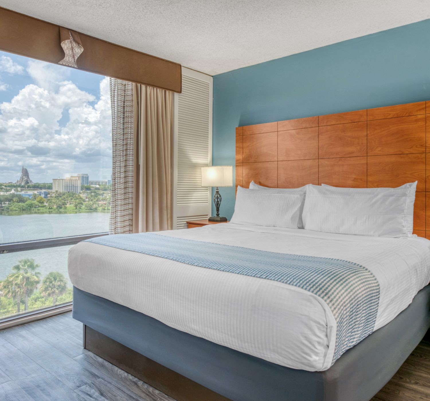 A hotel room with a large bed, bedside lamps, and a window view of a cityscape with a lake and clouds outside.
