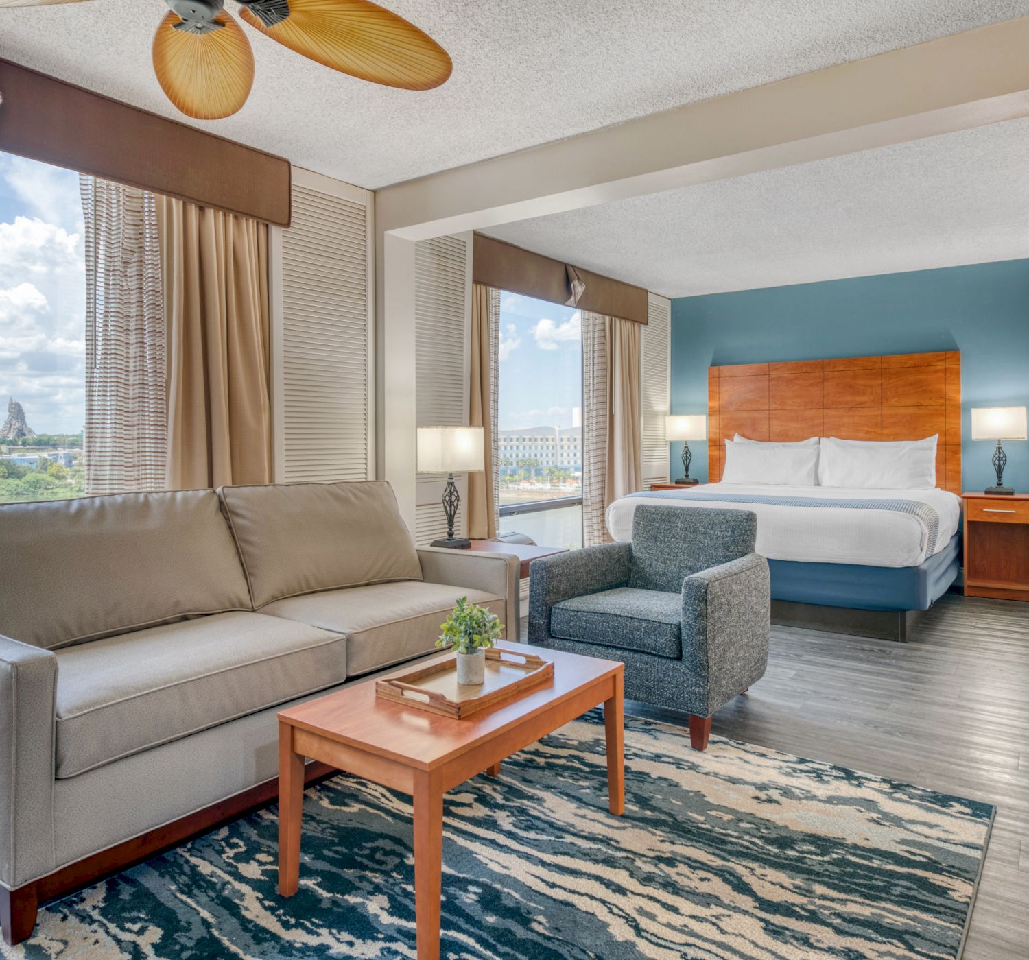 The image shows a hotel room with a sofa, armchair, bed, table, and window view of a cityscape.