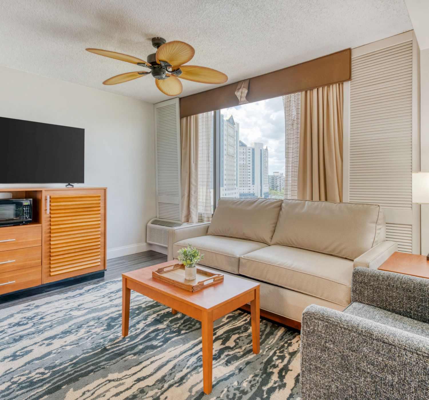 A modern living room with a sofa, chair, TV, microwave, coffee table, lamp, ceiling fan, and window view of a cityscape.