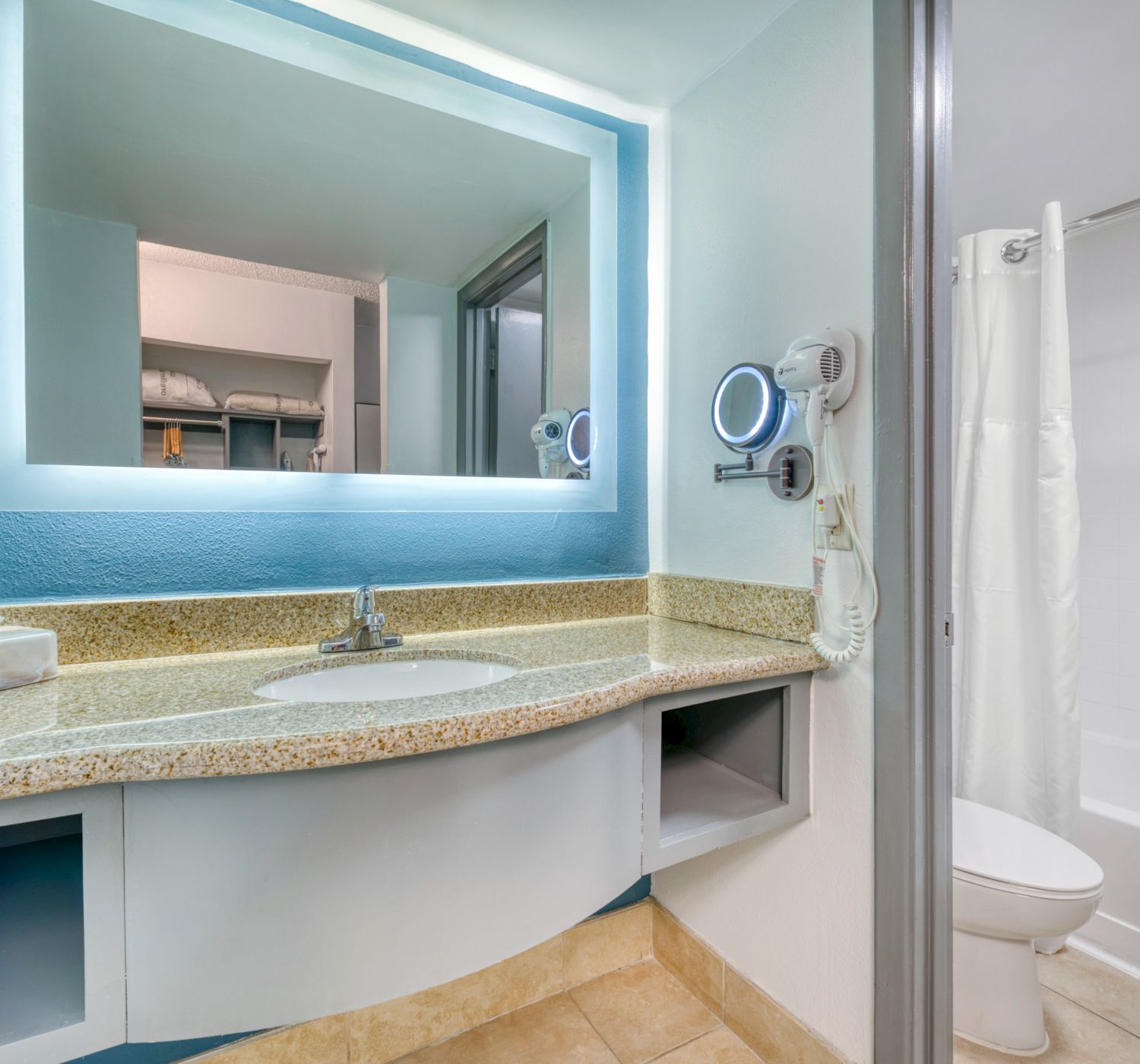 The image shows a bathroom with a lit mirror, sink, countertop, hairdryer, towels, and a shower with a curtain.