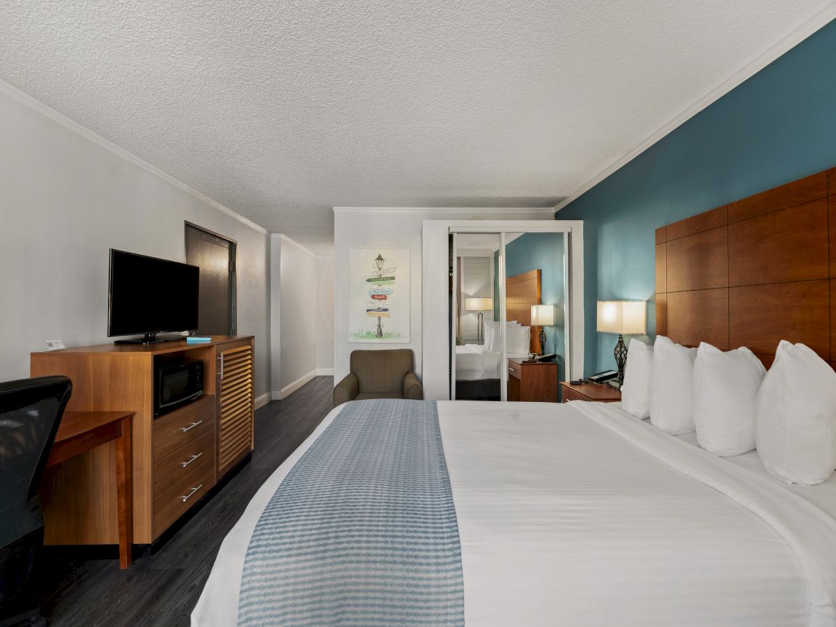 A hotel room with a king-sized bed, a TV on a dresser, a desk with a chair, and a bathroom in the background. The walls are white and teal.