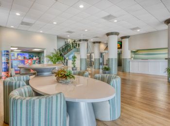 The image shows a spacious lobby with a round table, striped chairs, plants, and a staircase, creating a welcoming atmosphere.