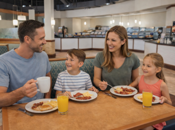 A family of four is enjoying breakfast in a restaurant, each with a plate of food and drinks, smiling and talking together.