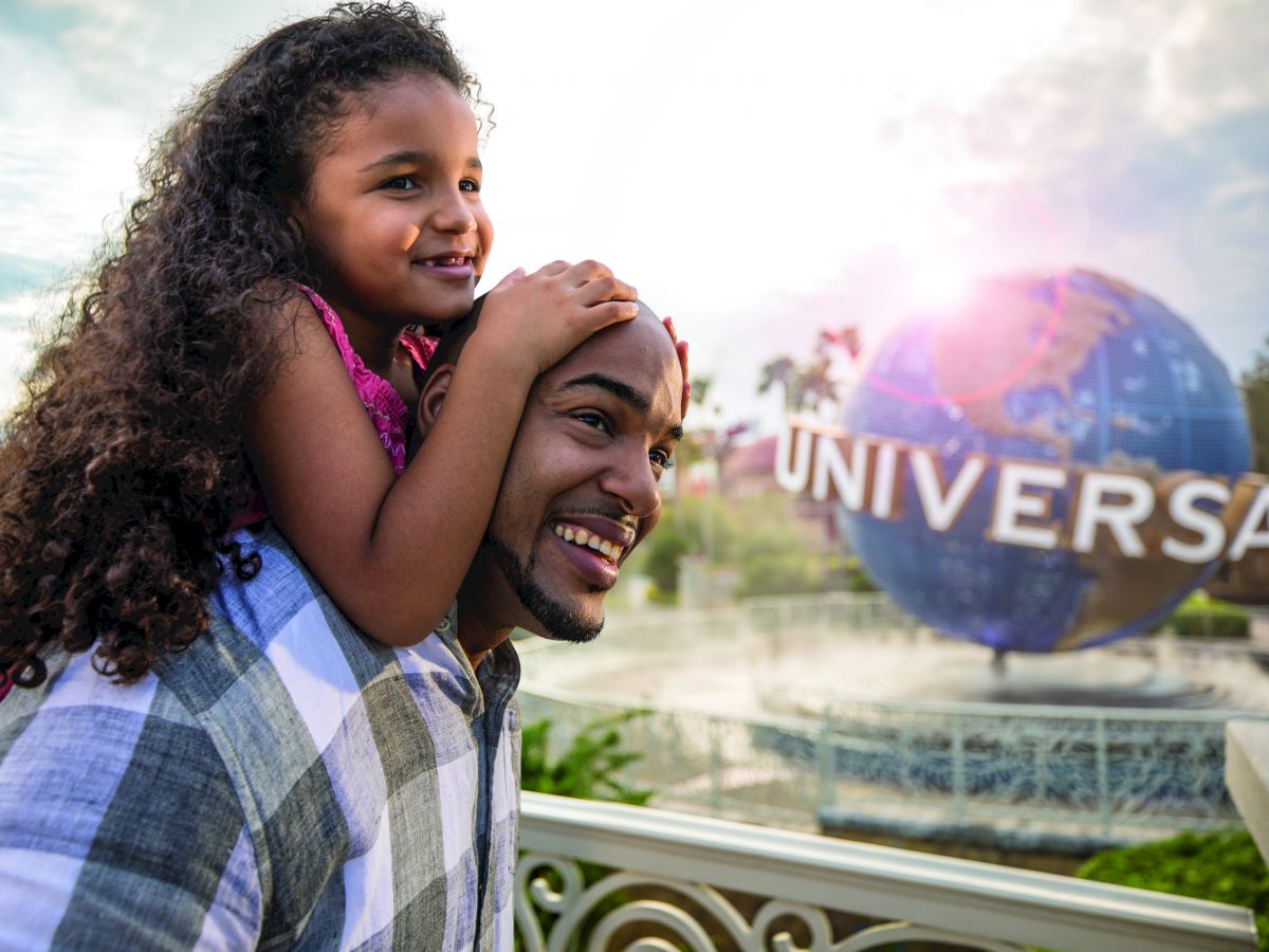 A person gives a young girl a piggyback ride near the iconic Universal globe, suggesting they are at a Universal theme park.