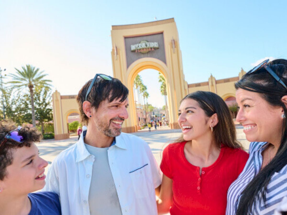 A group of people smiling in front of an entrance arch at a theme park, with palm trees and clear blue sky in the background.