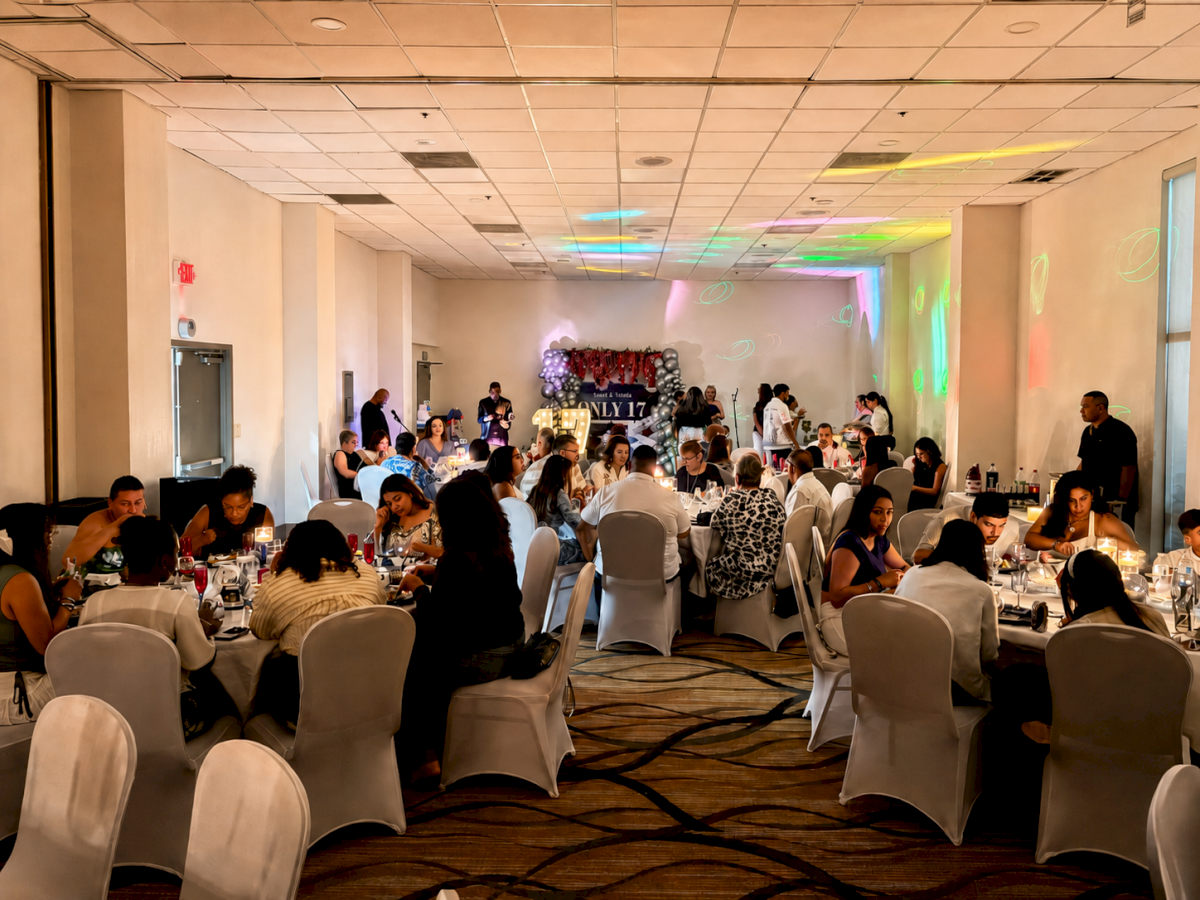 The image shows a decorated event hall with people seated at round tables, likely attending a social or formal gathering.