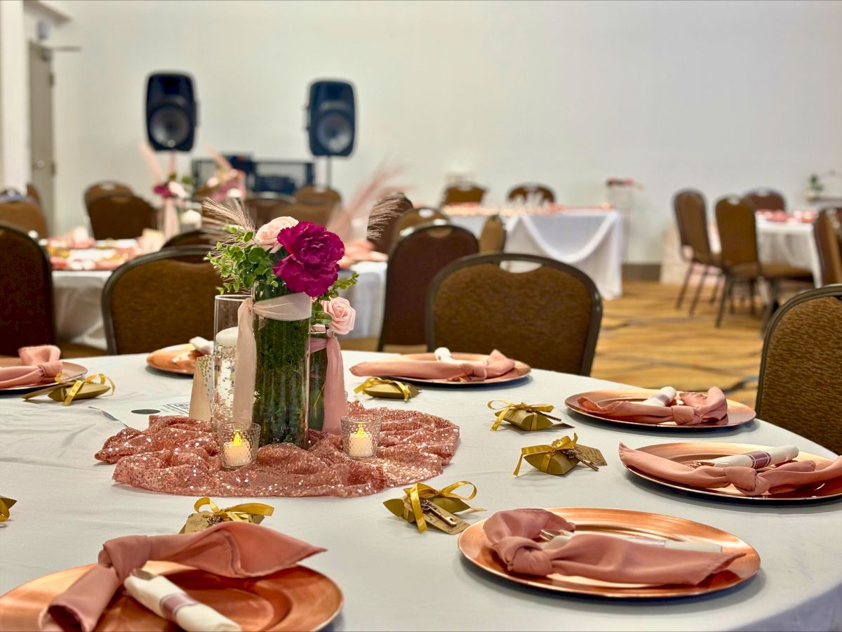 The image shows a decorated event venue with round tables set for dining, featuring floral centerpieces, candles, and neatly folded napkins.