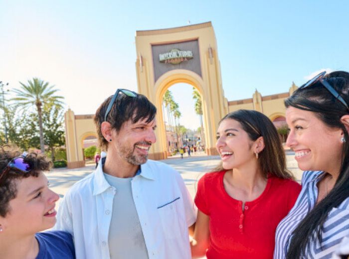 A group of four people smiling in front of an entrance with an archway and palm trees under a clear blue sky.