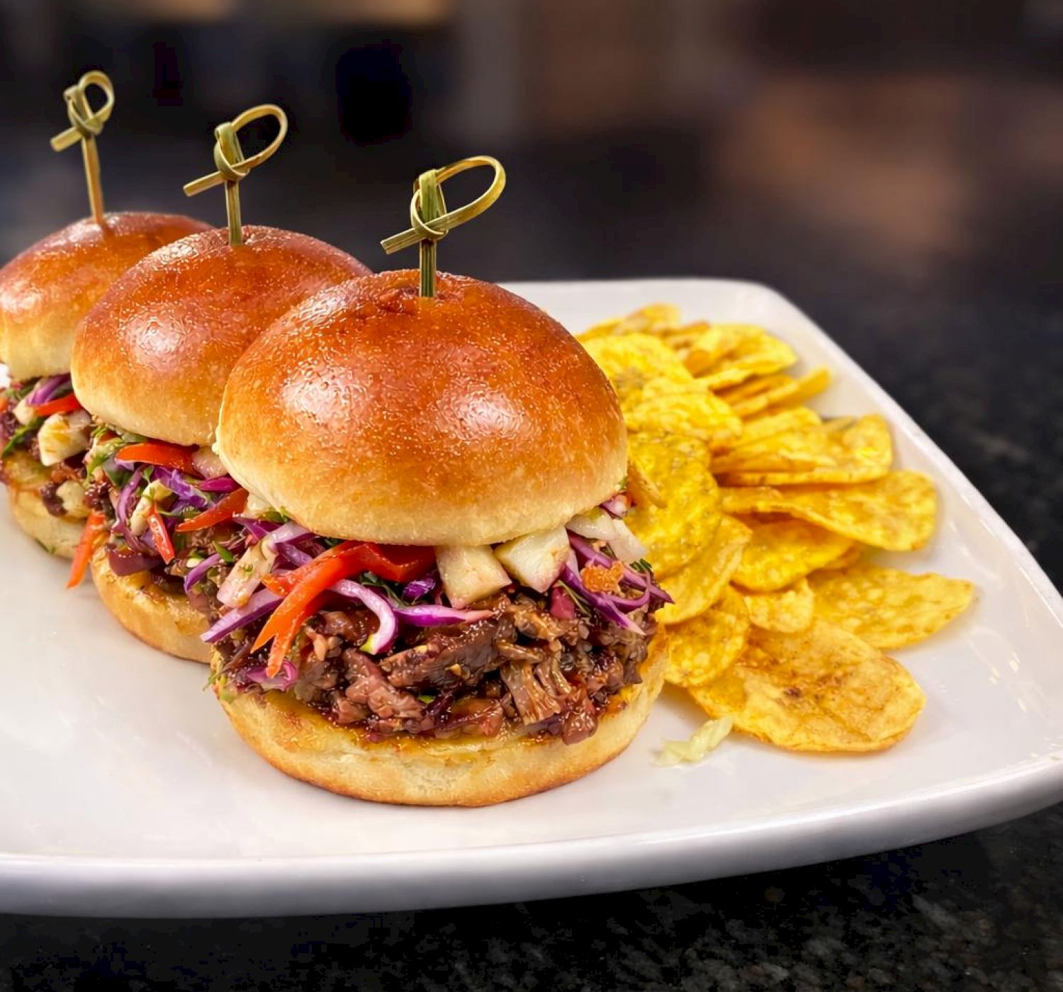 The image shows three sliders with coleslaw on a plate, accompanied by a side of plantain chips.