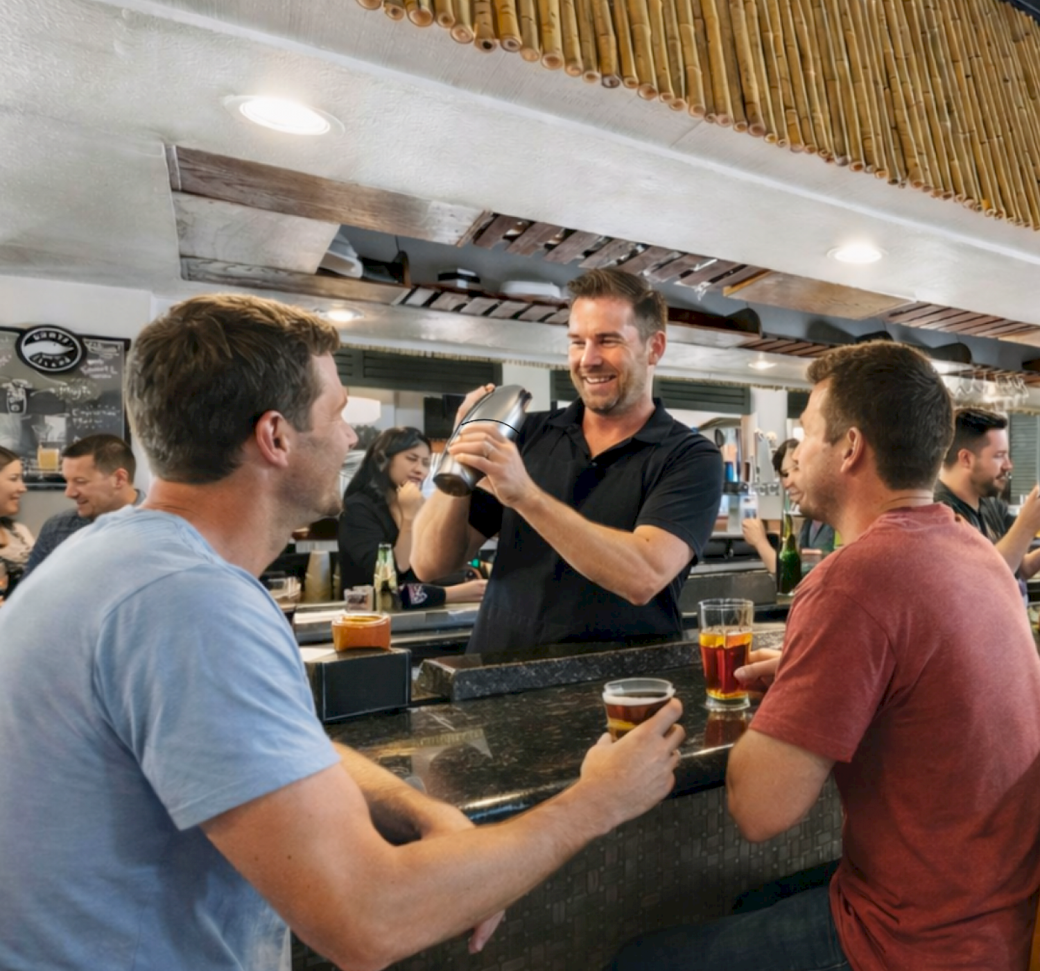 A bartender is mixing a drink while two patrons sit at the bar, each holding a glass, in a lively atmosphere.