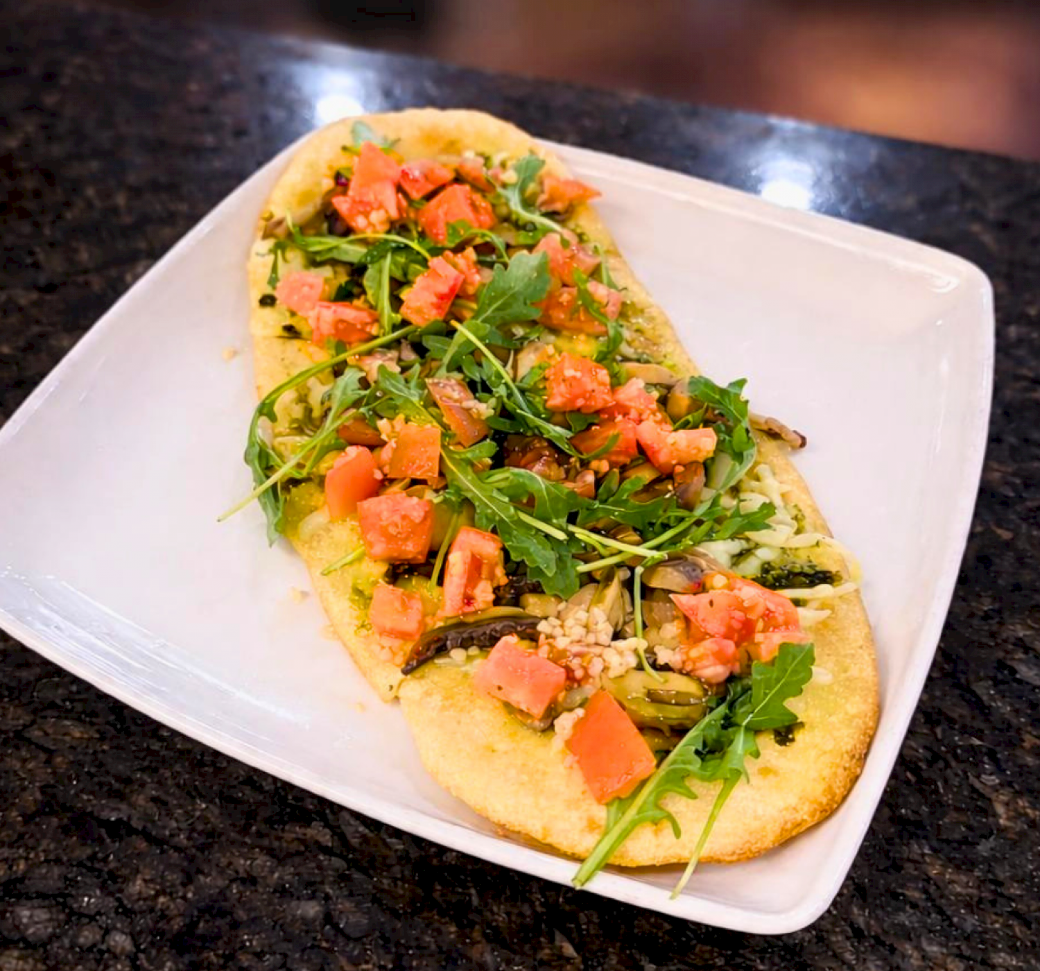 A flatbread topped with arugula, diced tomatoes, and what appears to be cheese sits on a white plate placed on a dark countertop.