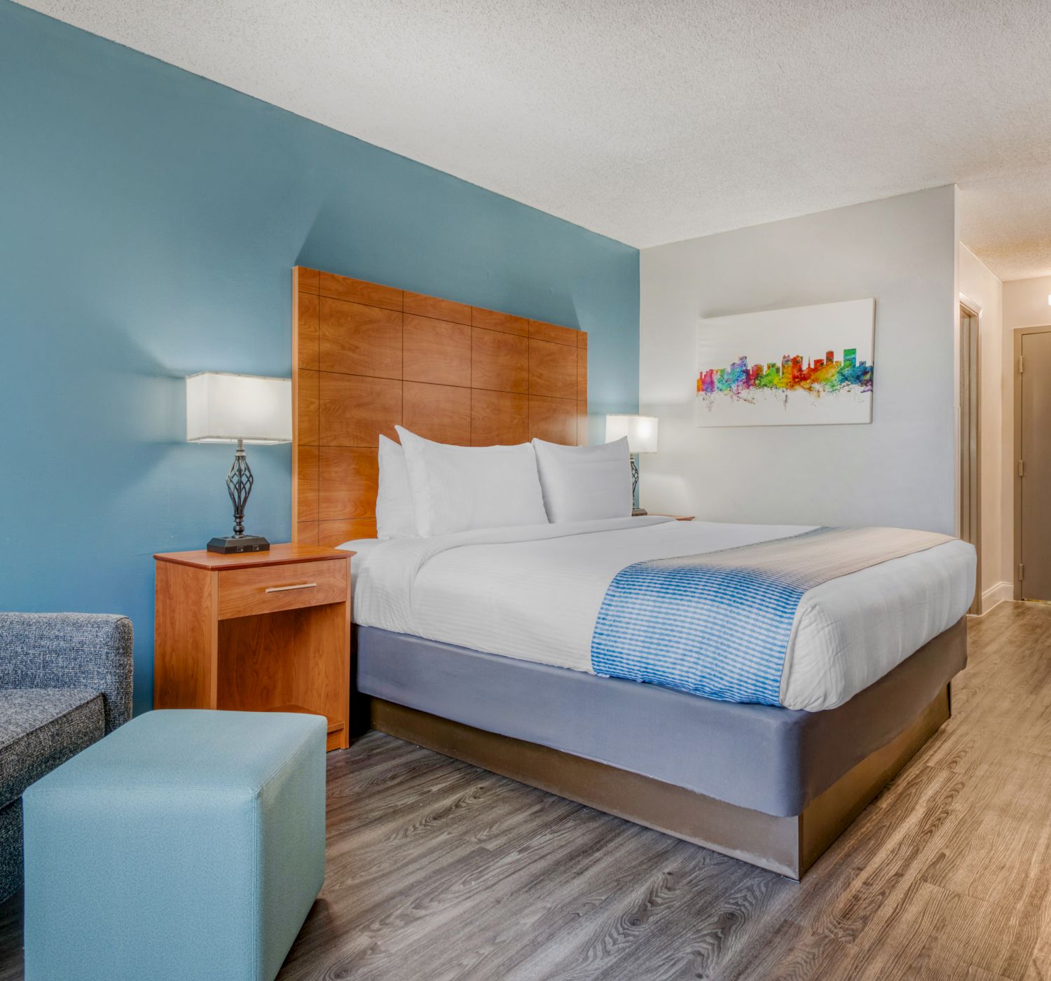 A modern hotel room with a large bed, blue accent wall, artwork, a nightstand, and a small seating area with a chair and ottoman.