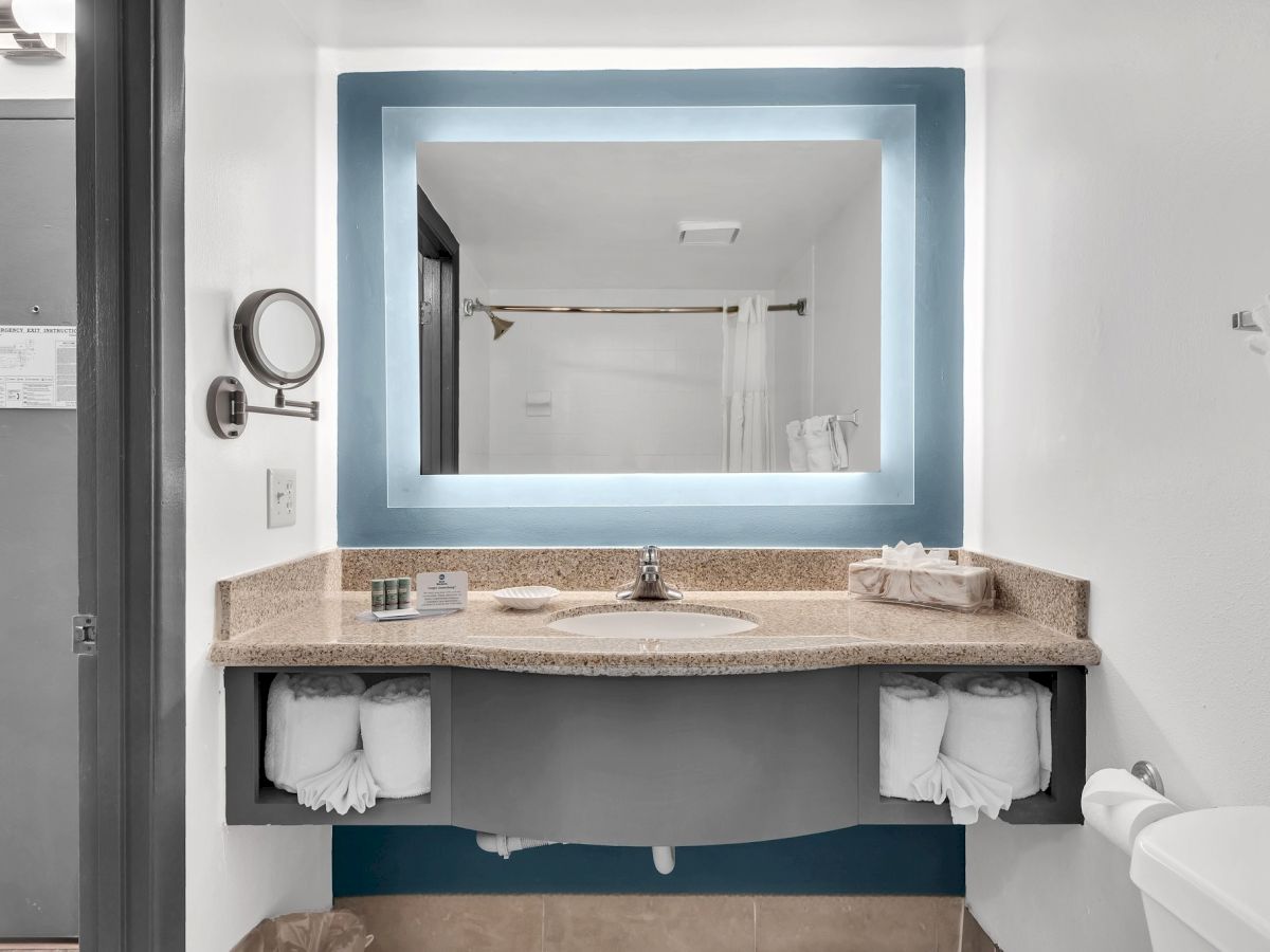 A bathroom with a large illuminated mirror, sink, towels, and a shower curtain in the reflection, creating a clean and modern look.