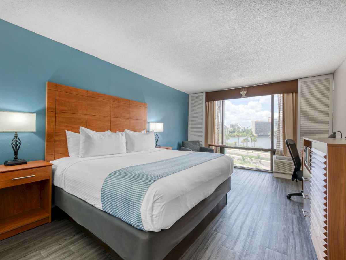 The image shows a modern hotel room with a large bed, side tables, lamps, a desk, and a window offering a view outside.