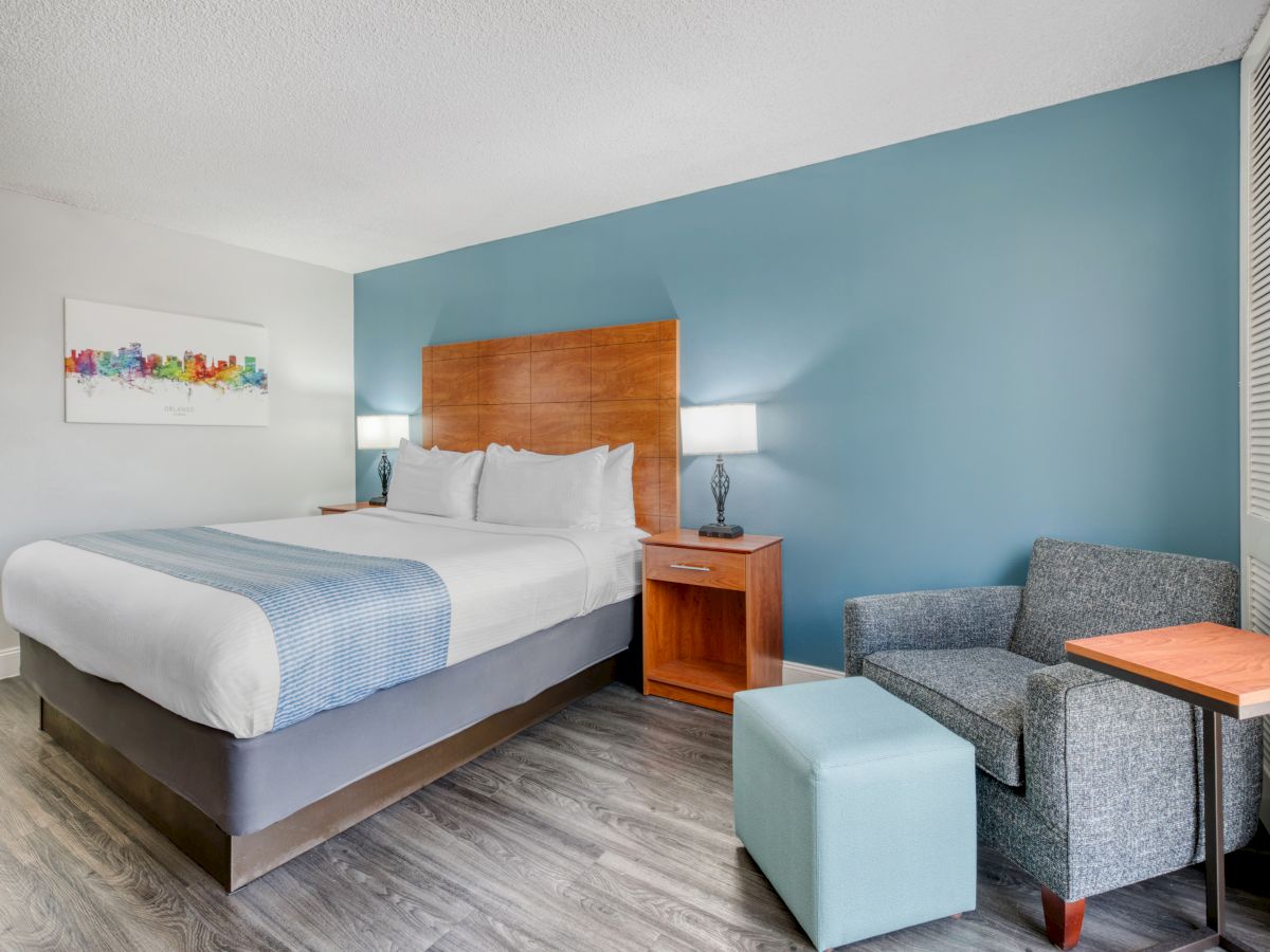A hotel room with a large bed, blue accent wall, armchair, and modern decor, featuring a wooden nightstand and lamps on either side.