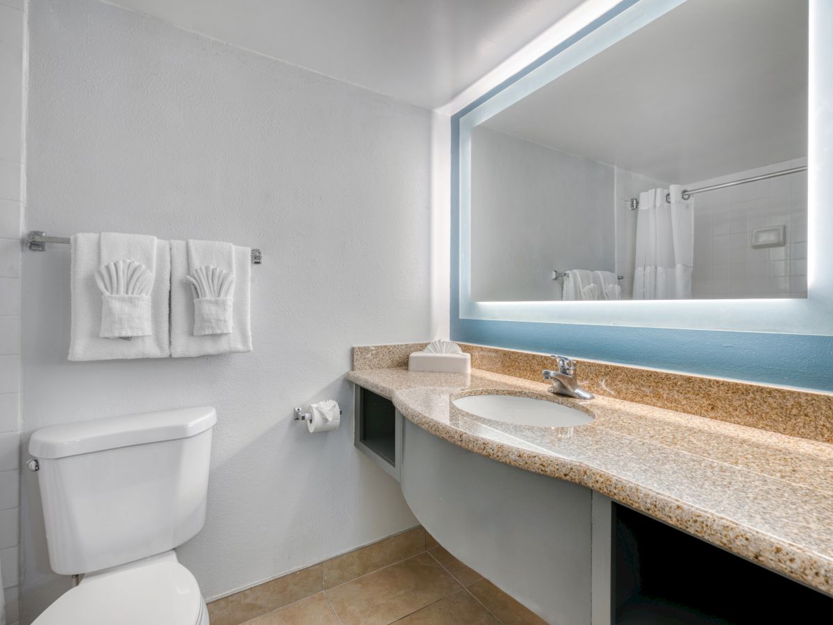 The image shows a bathroom with a granite countertop, a sink, a wall mirror, a toilet, and two towels neatly folded on a rack.