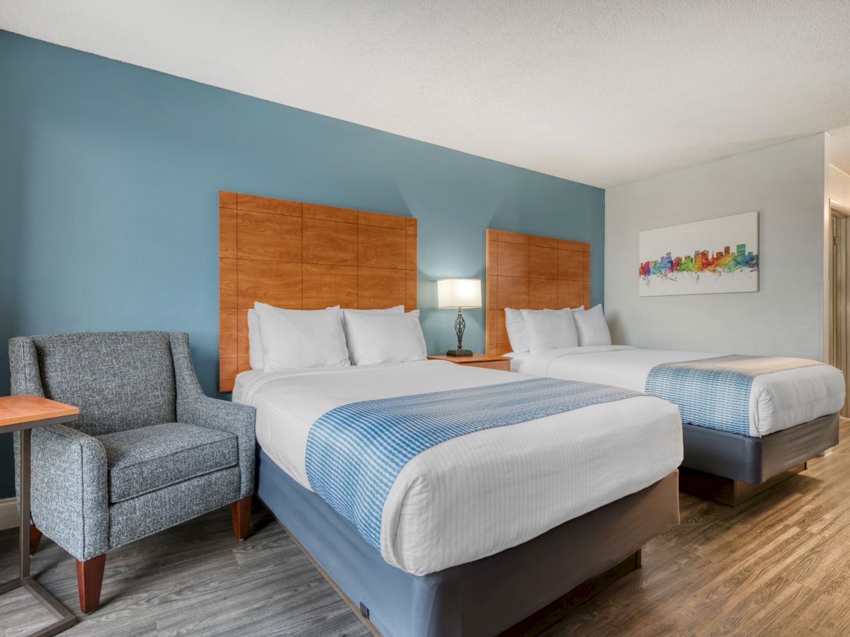 The image shows a hotel room with two beds, a chair, a lamp, and colorful artwork on the wall, featuring a modern design.