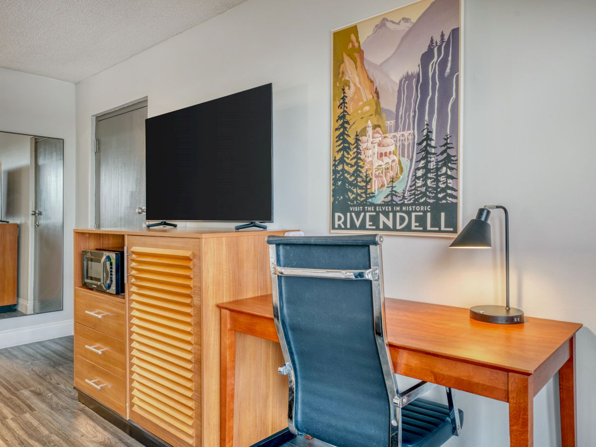 A modern room with a TV, wooden desk, chair, and Rivendell poster on the wall. A lamp is placed on the desk next to the chair.