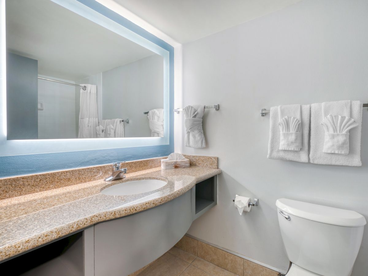 A clean bathroom with a large mirror, a countertop sink, a toilet, and neatly folded towels hanging on the wall.