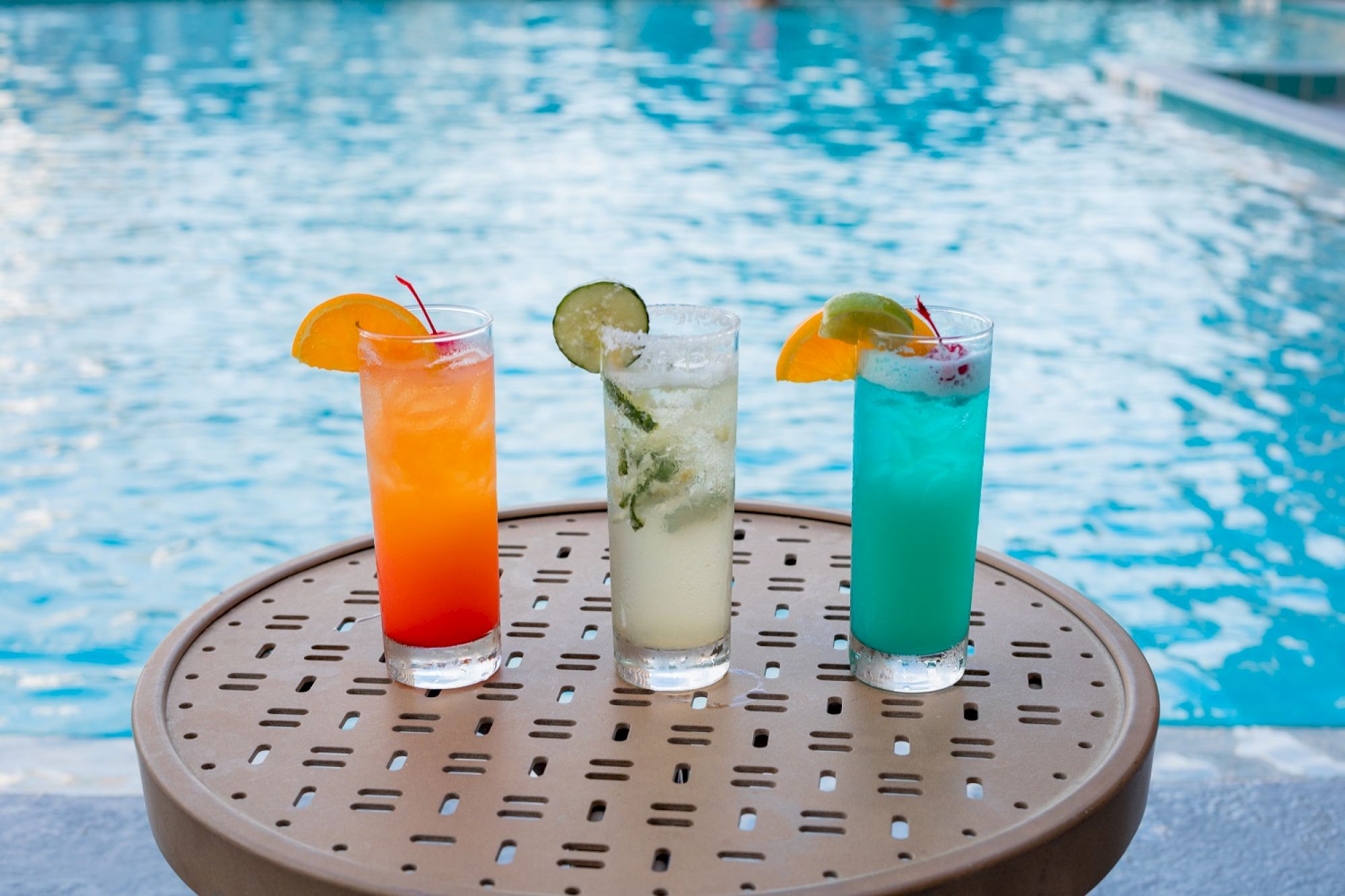 Three colorful cocktails garnished with fruit slices sit on a table by a pool.