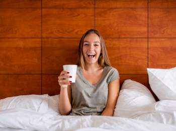 A person sitting in bed with white pillows and holding a coffee cup, smiling in a cozy room with wooden paneling behind.