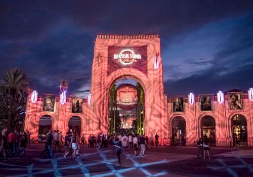 This image shows the entrance to Universal Studios at night, illuminated with red lighting and projections, with a crowd of people walking.
