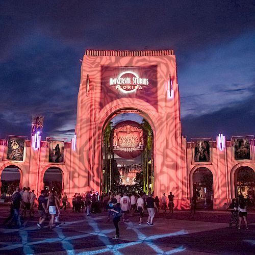 This image shows the entrance to Universal Studios at night, illuminated with red lighting and projections, with a crowd of people walking.