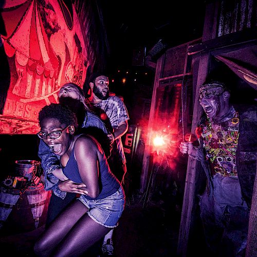 People are in a haunted house, appearing scared as an actor dressed as a zombie frightens them from a doorway with eerie lighting.