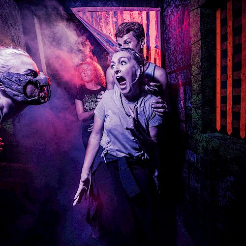 People are in a haunted house, reacting with fear to a costumed actor wearing a gas mask and makeup, under colorful, eerie lighting.