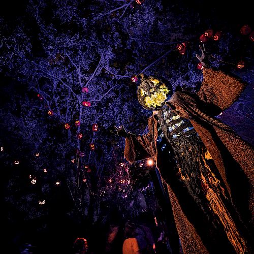 The image depicts a spooky, eerie scarecrow-like figure with a pumpkin head in a dark, forested area with glowing lights and night-time ambiance.