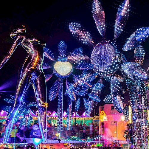 The image shows a vibrant nighttime festival scene with large illuminated figures, including a giant statue and glowing flowers under purple lighting.