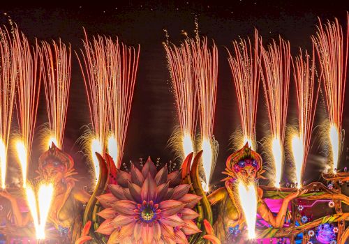A vibrant, large-scale music festival at night with colorful stage decorations, fireworks, and a crowd. "Orlando" is spelled out in the sky above the event.